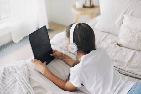 Photo for Adorable 12s pre-teen girl in wireless headphones lying on bed with digital tablet device. Young generation and modern tech overuse, gadget usage for fun, hobby and leisure on weekend at home - Royalty Free Image