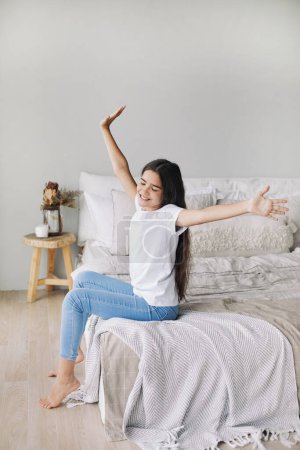 Photo for Pre-teen girl in casual clothes do stretching exercises seated on bed in cozy bedroom in the morning, raised her arms, feels refreshed after night sleep enjoy Sunday looks happy alone at home - Royalty Free Image