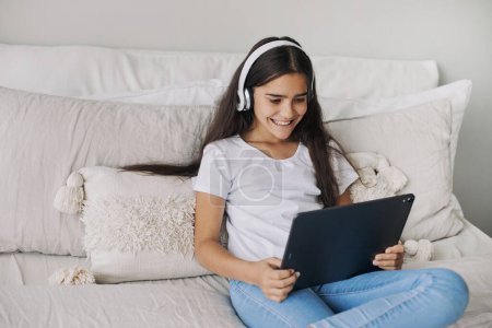 Téléchargez les photos : Attrayant pré-adolescent 12s fille porter des écouteurs sans fil à l'aide d'une tablette numérique moderne assis sur le lit dans une chambre confortable. I génération et utilisation des technologies pour le plaisir, l'éducation et les loisirs - en image libre de droit