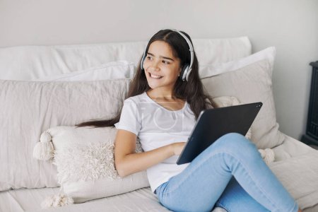 Foto de Atractivo pre-adolescente 12s niña usar auriculares inalámbricos utilizando tableta digital moderna sentada en la cama en el dormitorio acogedor. I generación y uso de tecnologías para la diversión, la educación y el ocio - Imagen libre de derechos