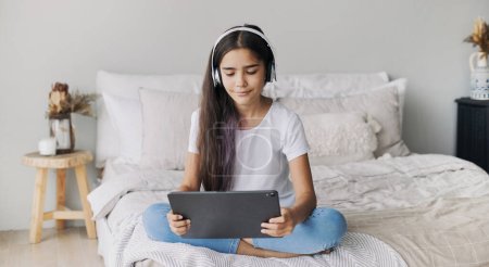Téléchargez les photos : Attrayant pré-adolescent 12s fille porter des écouteurs sans fil à l'aide d'une tablette numérique moderne assis sur le lit dans une chambre confortable. I génération et utilisation des technologies pour le plaisir, l'éducation et les loisirs - en image libre de droit