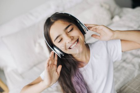 Téléchargez les photos : Jolie fille pré-adolescente écouter de la musique grâce à des écouteurs sans fil, vue rapprochée du visage. Utilisation de la technologie moderne, amusement, loisirs - en image libre de droit