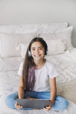 Foto de Atractivo pre-adolescente 12s niña usar auriculares inalámbricos utilizando tableta digital moderna sentada en la cama en el dormitorio acogedor. I generación y uso de tecnologías para la diversión, la educación y el ocio - Imagen libre de derechos