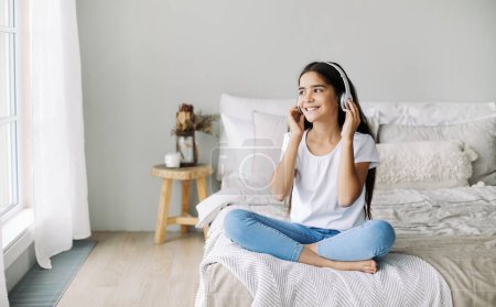 Foto de Bastante niña preadolescente escuchar música a través de auriculares inalámbricos, sentarse en la cama en casa disfrutar de la canción favorita se ve tranquilo en fin de semana en el dormitorio. Diversión, ocio - Imagen libre de derechos