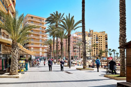 Téléchargez les photos : Torrevieja, Espagne-14 mars 2024 : Les touristes se promènent le long de la promenade en bord de mer avec une rue bordée de palmiers avec un café, des restaurants pendant la journée chaude ensoleillée de printemps. Destinations de voyage. Costa Blanca - en image libre de droit
