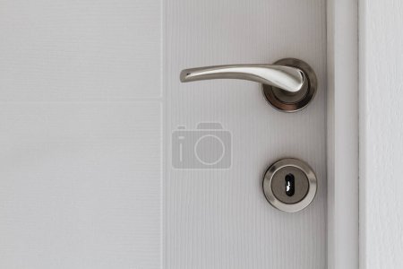 Poignée de porte argentée sur une porte blanche ouverte, révélant une partie d'un mur gris.