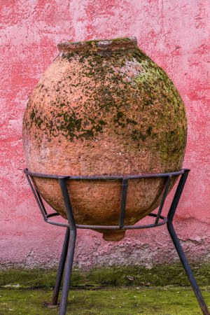 Antique ceramic jar on a forged pedestal against an aged pink wall.