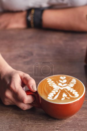 Photo for Person gently holding a red cup of coffee with an intricate, symmetrical floral latte art design on the surface of the coffee. - Royalty Free Image