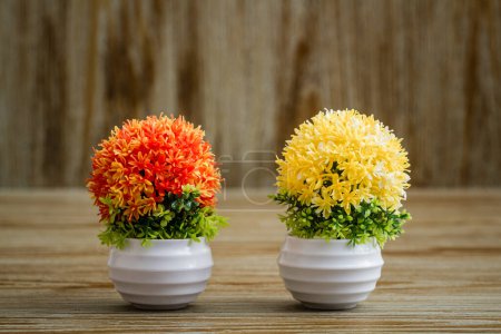 Photo for Vibrant, artificial orange,yellow flower arrangements placed on a rustic wooden surface, potted in a white pot that contrasts nicely with the colorful blossoms. - Royalty Free Image