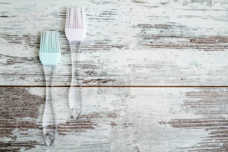 Two silicone basting brushes with transparent handles, one with a pink head and the other with a light blue head, placed on a rustic wooden white surface and with copy space.
