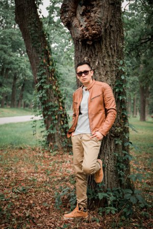 Téléchargez les photos : Un jeune homme beau se tient sous un arbre dans un parc d'automne - en image libre de droit
