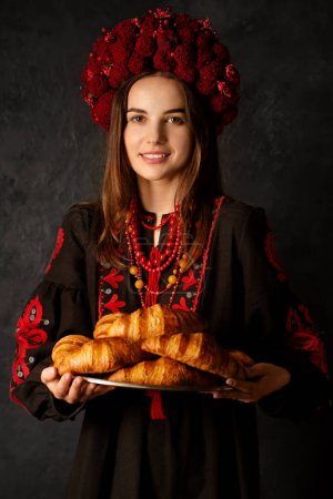 Photo for Ukrainian woman in national dress holds a tray with rosy croissants on a dark background - Royalty Free Image