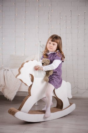 Una niña monta en un caballo de cartón de juguete en la habitación de los niños