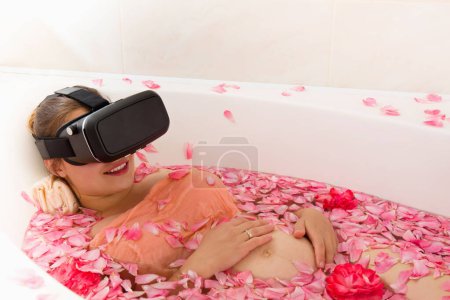 A pregnant woman in 3D virtual reality glasses rests in a bath with rose petals, on a light background