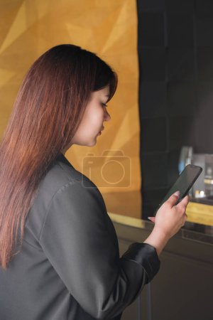 Téléchargez les photos : Belle jeune femme confiante en costume de bureau noir regardant smartphone en intérieur doré, photo de profil - en image libre de droit