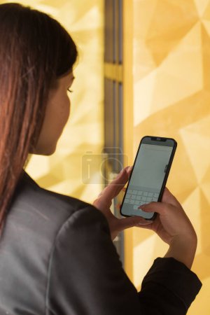 Téléchargez les photos : Belle jeune femme confiante en costume de bureau noir regardant smartphone en intérieur doré, photo de dos - en image libre de droit