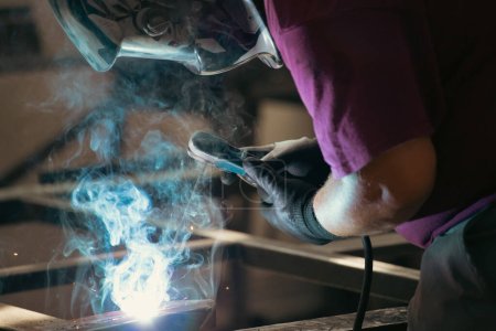 Téléchargez les photos : Handyman performing welding and grinding at his workplace in the workshop, while the sparks "fly" all around him. He is wearing a protective helmet and equipment. - en image libre de droit