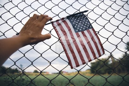 Primer plano de una bandera americana en la mano unida a una cerca de eslabones de cadena. Inmigración americana y concepto de crisis de refugiados de Estados Unidos