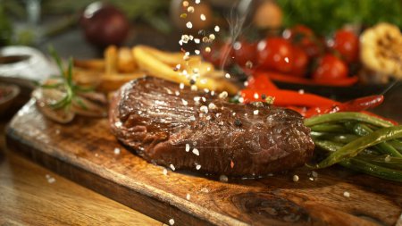 Téléchargez les photos : Gros plan sur la chute de sel sur un délicieux steak de boeuf dans la cuisine. - en image libre de droit