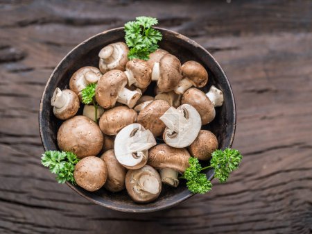 Photo for Brown colored common mushrooms in wooden bowl on wooden table with herbs. Top view. - Royalty Free Image