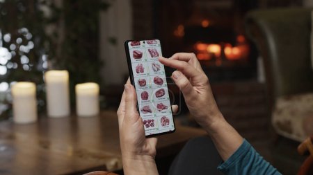 Téléchargez les photos : Commande de nourriture à l'aide d'un smartphone à la maison. Une femme sélectionne la viande et le poisson dans une boutique en ligne à l'aide d'une application sur un smartphone. Aménagement du soir avec un feu brûlant dans la cheminée. - en image libre de droit