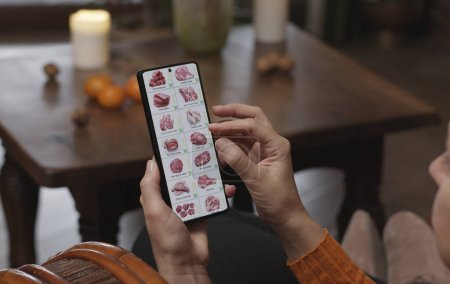 Photo for Ordering food using a smartphone at home. A woman selects meat and fish in an online store using an application on a smartphone. Home evening furnishings with a burning fire in the fireplace. - Royalty Free Image