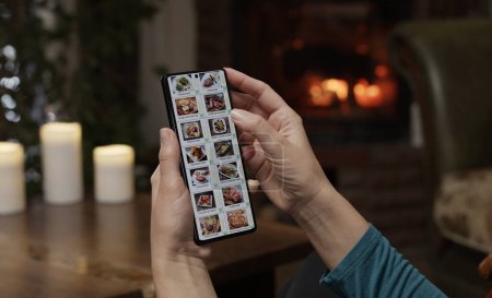 Téléchargez les photos : Ordering food using a smartphone at home. A woman selects restaurant food in the internet menu of a gourmet restaurant using an application on a smartphone. Home evening furnishings with a burning fire in the fireplace. - en image libre de droit