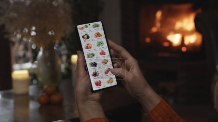 Foto de Pedir comida usando un smartphone en casa. Una mujer selecciona verduras en una tienda en línea utilizando una aplicación en un teléfono inteligente. Muebles de casa por la noche con una chimenea en llamas en la chimenea. - Imagen libre de derechos