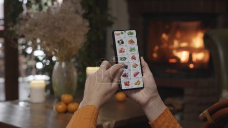 Téléchargez les photos : Commande de nourriture à l'aide d'un smartphone à la maison. Une femme sélectionne des légumes dans une boutique en ligne à l'aide d'une application sur un smartphone. Aménagement du soir avec un feu brûlant dans la cheminée. - en image libre de droit