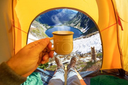 Téléchargez les photos : Coupe de boisson chaude dans la main et magnifique lac de montagne à travers l'entrée ouverte de la tente. La beauté d'une randonnée romantique et camping accompagné d'un chien. - en image libre de droit
