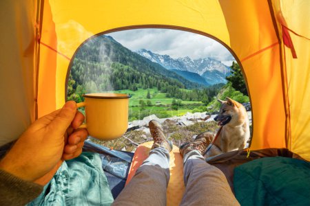 Photo for Cup of hot drink in the hand and wonderful view of snowy mountain tops through the open entrance of the tent. The beauty of a romantic hike and camping accompanied by a dog. - Royalty Free Image