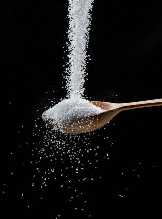 Photo for White refined sugar crystals falling down into the wooden spoon at black background. - Royalty Free Image