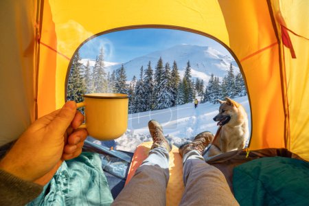 Photo for Hot drink in the hand and wonderful view of snowy forest through the open entrance to the tent. The beauty of a romantic hike and camping accompanied by a dog. - Royalty Free Image