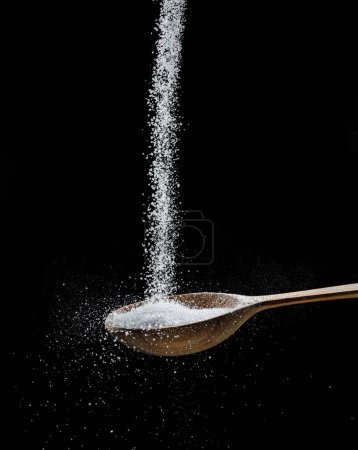 Foto de Cristales de azúcar refinados blancos cayendo en la cuchara de madera en el fondo negro. - Imagen libre de derechos