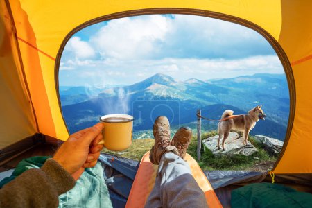 Téléchargez les photos : Coupe de boisson chaude dans la main et vue magnifique sur les sommets de montagne à travers l'entrée ouverte de la tente. La beauté d'une randonnée romantique et camping accompagné d'un chien. - en image libre de droit