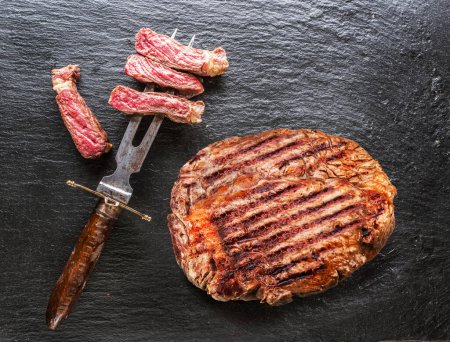 Photo for Grilled delicious ribeye steak and some pieces of steak on fork on grey background. Top view. - Royalty Free Image