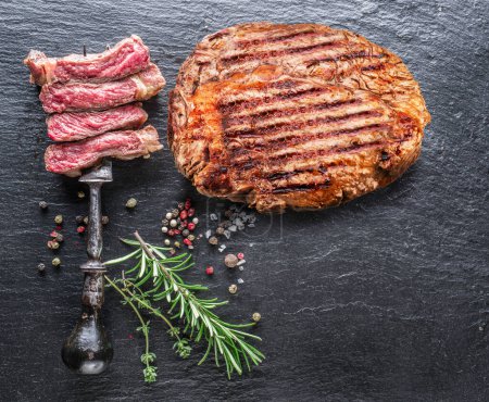 Photo for Grilled delicious ribeye steak, spices and pieces of medium steak on fork on grey background. Top view. - Royalty Free Image