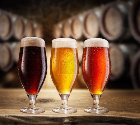 Photo for Three type of beer with foam head on wooden table. Blurred brewery cellar at the background. - Royalty Free Image