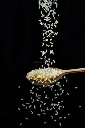 Photo for White rice grains falling down into the spoon at black background. Popular food and main ingredient of risotto and pilau. - Royalty Free Image