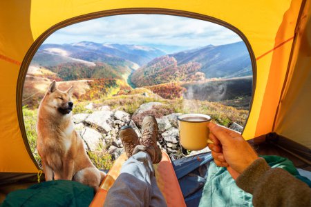 Photo for Cup of hot drink in the hand and wonderful view of autumn mountain tops through the open entrance of the tent. The beauty of a romantic hike and camping accompanied by a dog. - Royalty Free Image