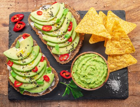 Avocado toasts - bread with avocado slices, pieces of red pepper and sesame  on black slate serving plate.