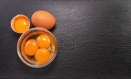 Photo for Brown chicken egg and egg yolks in glass bowl on natural stone black board. Flat lay. - Royalty Free Image