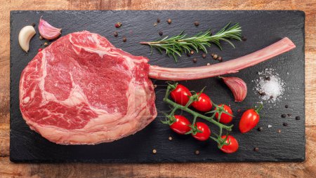 Photo for Raw rib steak with bone or tomahawk steak with seasonings on slate serving plate. Flat lay. - Royalty Free Image
