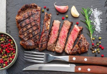 Photo for Grilled  delicious ribeye steak slices and some seasonings on black slate serving plate. Flat lay. - Royalty Free Image