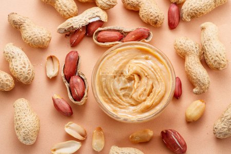 Photo for Bowl of peanut butter and peanuts around it on beige background. Top view. - Royalty Free Image
