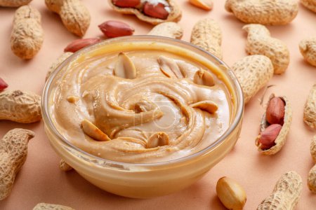 Bowl of peanut butter and peanuts around it on beige background. 