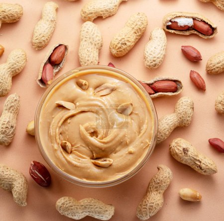 Photo for Bowl of peanut butter and peanuts around it on beige background. Top view. - Royalty Free Image