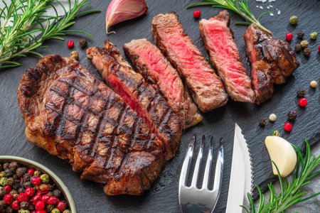 Foto de Grilled  delicious ribeye steak slices and some seasonings on black slate serving plate. Flat lay. - Imagen libre de derechos