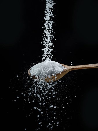 Foto de Cristales de sal comestibles cayendo en la cuchara de madera en el fondo negro. - Imagen libre de derechos