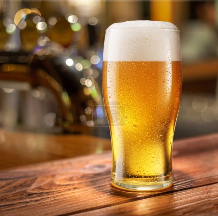 Glass of chilled beer on wooden bar table top and blurred bar interior at the background.